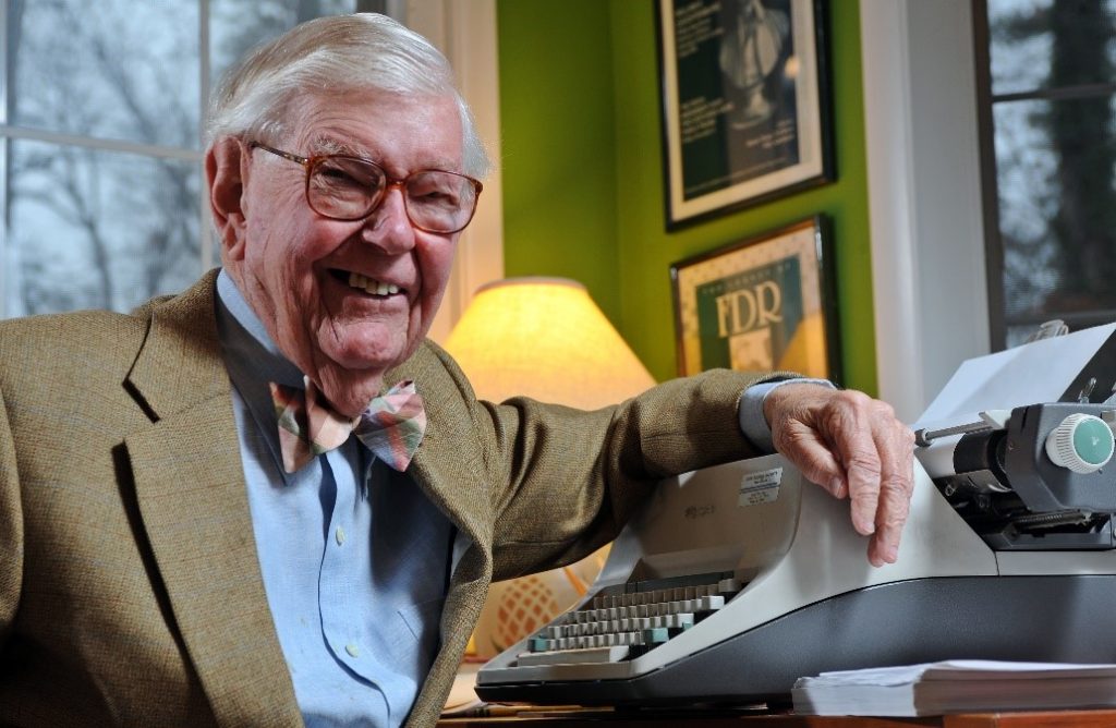 Professor Bill Leuchtenburg, smiles widely while wearing a brown suit, blue shirt and a bowtie. He leans his left arm over a typewriter at a desk.